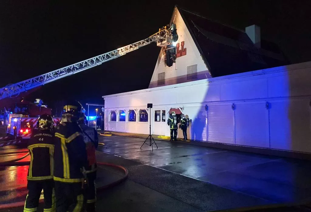 Des pompiers en tenue de protection interviennent sur un incendie nocturne à Nantes, en Loire-Atlantique. Un camion-échelle s&#039;étend jusqu&#039;au toit, où un pompier travaille avec diligence. Des lampes de secours d&#039;Actu44.fr éclairent la scène et des lances sont visibles sur le sol humide.