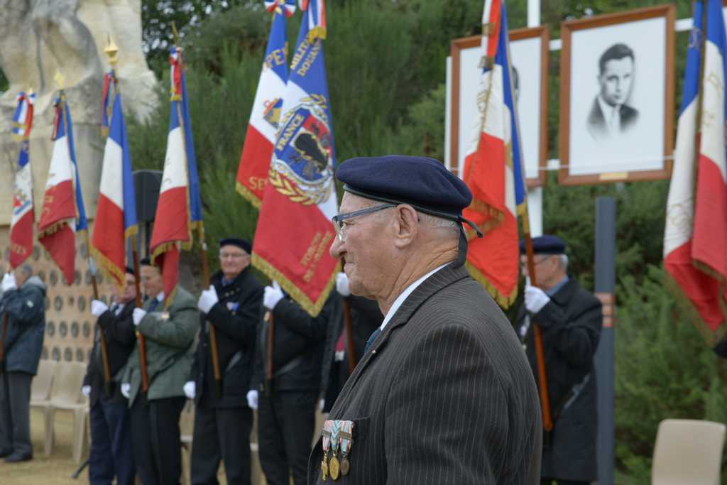 Un homme âgé portant un béret se tient debout lors d&#039;une cérémonie solennelle à Nantes, paré de médailles. Derrière lui, plusieurs personnes brandissent des drapeaux français. Des portraits photographiques encadrés sont exposés sur des chevalets, dans un décor de verdure au cœur de la Loire-Atlantique.