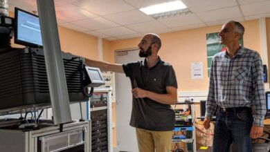 Dans un laboratoire technologique de Nantes, deux hommes sont plongés dans leur travail. L'un pointe du doigt un écran d'ordinateur perché sur une haute pile d'équipements, tandis que l'autre observe attentivement. Des murs beiges et un éclairage fluorescent les entourent, avec divers câbles et appareils éparpillés un peu partout.