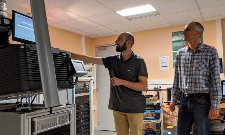 Dans un laboratoire technologique de Nantes, deux hommes sont plongés dans leur travail. L'un pointe du doigt un écran d'ordinateur perché sur une haute pile d'équipements, tandis que l'autre observe attentivement. Des murs beiges et un éclairage fluorescent les entourent, avec divers câbles et appareils éparpillés un peu partout.