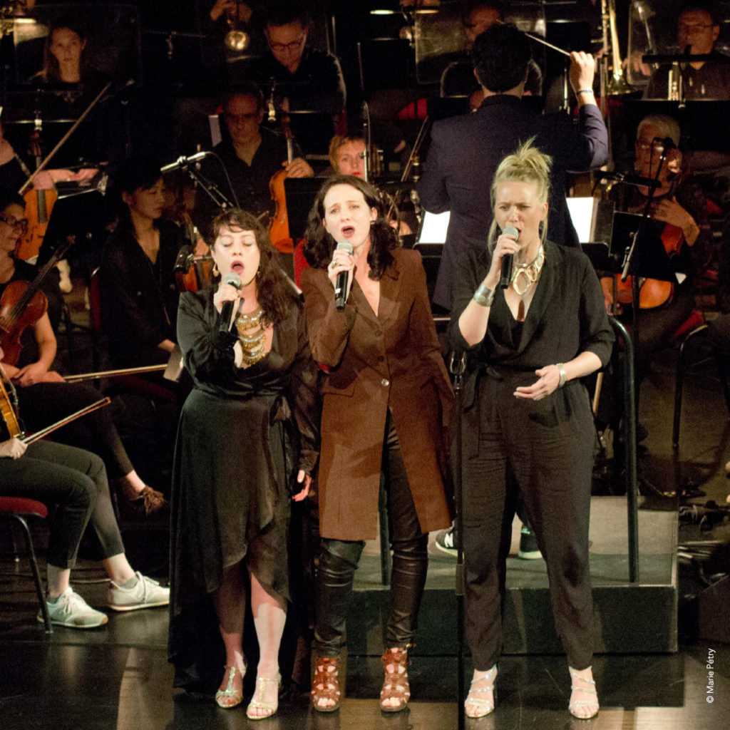 Trois femmes chantent sur scène, munies de micros, devant un orchestre dans une ambiance tamisée. Vêtues de tenues sombres, chacune affiche un style unique. Derrière elles, le chef d&#039;orchestre et les musiciens de Loire-Atlantique jouent en harmonie, créant une atmosphère enchanteresse.