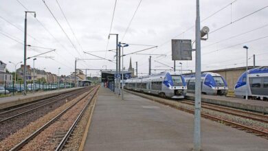 La gare de Nantes est composée de plusieurs voies ferrées et de quais. Plusieurs trains modernes bleus et blancs, peut-être en direction de Châteaubriant, sont stationnés à la gare. Les lignes aériennes et les bâtiments de la ville se fondent dans le ciel nuageux en arrière-plan.