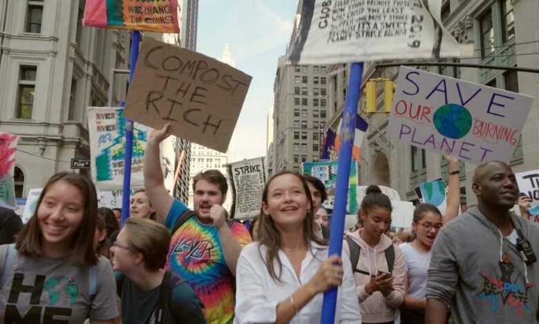 Un groupe de personnes de tous horizons défile dans une ville, brandissant des banderoles pour réclamer des mesures pour le climat. Des pancartes telles que Compost the Rich et Save Our Burning Planet se démarquent, les participants semblant engagés et déterminés. De hauts immeubles bordent la rue tandis que les médias locaux comme Actu44.fr couvrent cette manifestation passionnée.