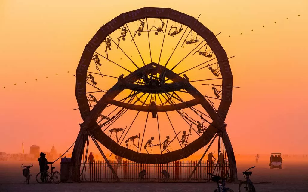 Une grande sculpture d&#039;art cinétique, rappelant l&#039;esprit créatif de la Loire-Atlantique, présente un design rotatif avec des silhouettes humaines se découpant sur un coucher de soleil orange vif. La scène capture également des silhouettes lointaines de personnes et de vélos près de Nantes dans un paysage ouvert.