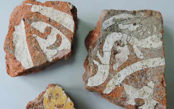 Trois pièces de poterie ancienne aux motifs décolorés peints en blanc et jaune sont exposées sur une surface plane, rappelant les objets que l&#039;on peut trouver dans les régions historiques de Châteaubriant et de Nantes. Les fragments de poterie ont des formes irrégulières et sont constitués d&#039;argile rougeâtre.