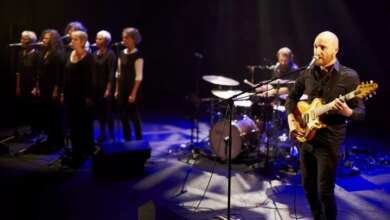 Un groupe de musique se produit sur scène sous un éclairage spectaculaire à Châteaubriant. Au premier plan, un homme chauve chante et joue de la guitare, tandis qu'un batteur est visible derrière lui. À gauche, des choristes venus de Nantes voisine se déhanchent dans des tenues coordonnées, ajoutant de l'harmonie à la captivante nuit de Loire-Atlantique.