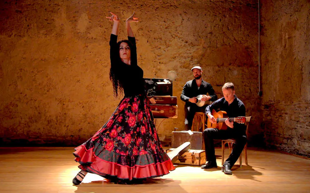 Une danseuse de flamenco vêtue d&#039;une robe fleurie rouge et noire vibrante se produit les bras levés au cœur de Nantes, tandis que deux musiciens, l&#039;un jouant de la guitare et l&#039;autre d&#039;un instrument à cordes, l&#039;accompagnent contre un mur rustique et texturé.