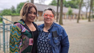 Deux femmes se tiennent dehors par une journée nuageuse en Loire-Atlantique. L'une porte une veste à motifs colorés, l'autre une veste en jean et des lunettes rouges. Un chemin de gravier et des arbres sont en arrière-plan. Toutes deux sourient à l'objectif, capturant un moment qui pourrait orner les pages d'Actu44.fr sur la vie animée de Nantes.