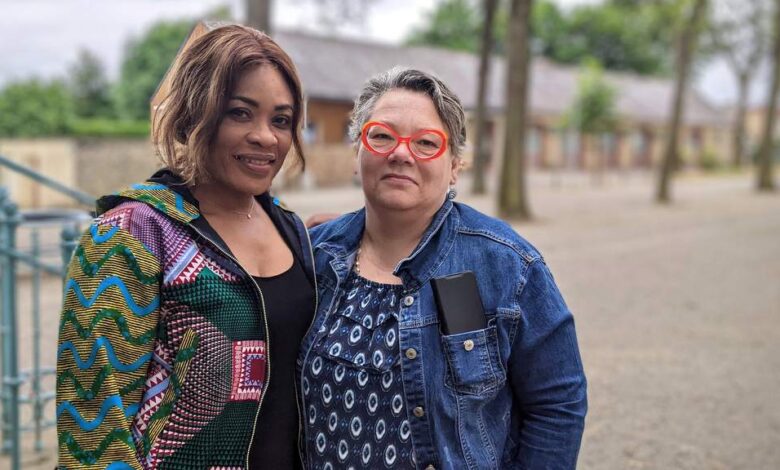 Deux femmes se tiennent dehors par une journée nuageuse en Loire-Atlantique. L'une porte une veste à motifs colorés, l'autre une veste en jean et des lunettes rouges. Un chemin de gravier et des arbres sont en arrière-plan. Toutes deux sourient à l'objectif, capturant un moment qui pourrait orner les pages d'Actu44.fr sur la vie animée de Nantes.