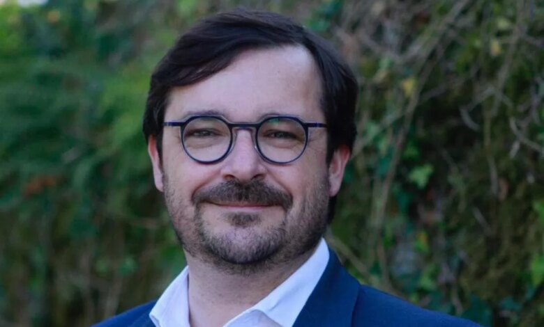 Un homme aux cheveux noirs et à la barbe, portant des lunettes et un costume bleu avec une chemise blanche, se tient souriant devant la verdure luxuriante de Châteaubriant, capturant l'essence vibrante de la Loire-Atlantique.