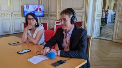 Deux personnes à une table avec un micro. L'homme à droite, portant un casque et un costume, parle au micro, évoquant les derniers événements à Nantes. La femme à gauche, en haut blanc et lunettes, est assise pensivement tandis que la télévision en arrière-plan diffuse des images d'actualité de la Loire-Atlantique.