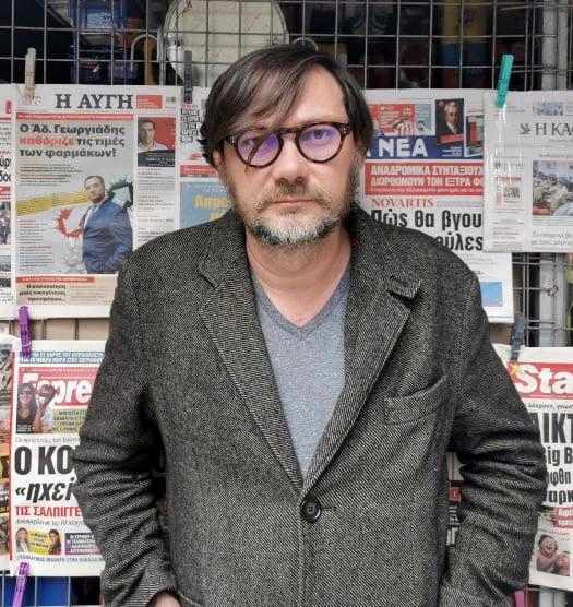 Un homme portant des lunettes et un blazer gris se tient devant un kiosque à journaux à Nantes, exposant divers journaux.