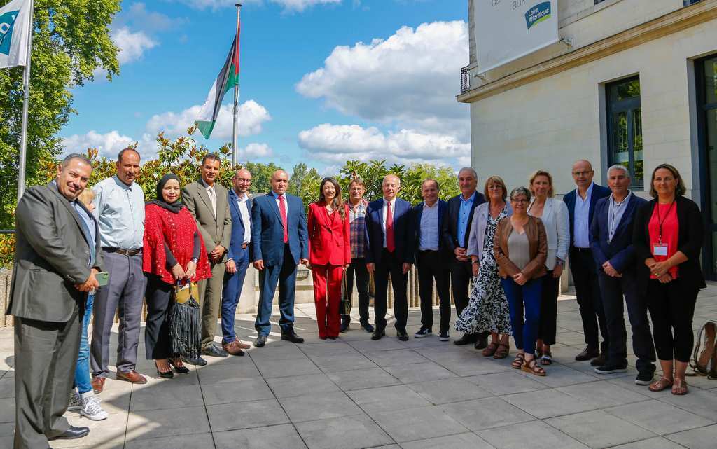 Un groupe de personnes se tient en rang sur une terrasse extérieure, vêtues de tenues de soirée. Avec le drapeau de Châteaubriant flottant derrière eux et la beauté des arbres de Loire-Atlantique sous un ciel partiellement nuageux, hommes et femmes dégagent un air d&#039;élégance.