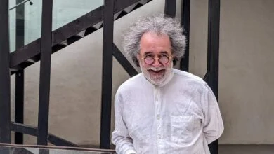 Un homme aux cheveux gris bouclés et aux lunettes rondes sourit tout en portant une chemise blanche. Il se tient devant un escalier en métal noir et un mur beige, rappelant l'architecture pleine de charme de Nantes.