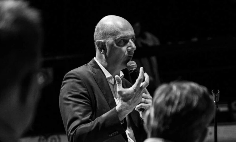 Sur une photo en noir et blanc, un homme en costume parle dans un micro, gesticulant avec passion. Capturé à Châteaubriant, des silhouettes floues remplissent le premier plan tandis qu'il partage son message avec la foule, faisant écho à l'actualité d'Actu44.fr.