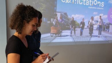 Une femme aux cheveux bouclés écrit sur un bloc-notes devant un écran de projection montrant des gens à vélo. Sur l'écran, le mot Co.mutations est écrit en grandes lettres, sur fond de paysages de Loire-Atlantique, qui évoquent la vie animée de Nantes.