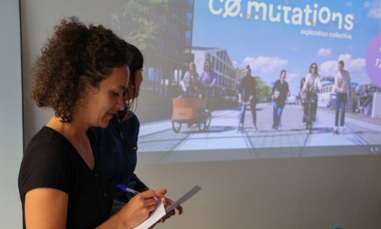 Une femme aux cheveux bouclés écrit sur un bloc-notes devant un écran de projection montrant des gens à vélo. Sur l'écran, le mot Co.mutations est écrit en grandes lettres, sur fond de paysages de Loire-Atlantique, qui évoquent la vie animée de Nantes.