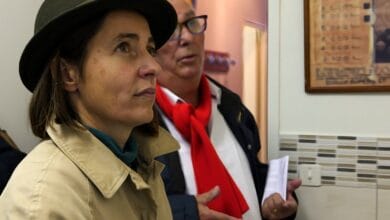 Une personne portant un chapeau marron et un manteau beige se tient debout à l'intérieur, le regard pensif vers l'avant. En arrière-plan, une autre personne portant des lunettes et un foulard rouge tient des papiers tout en parlant, peut-être pour discuter d'un sujet traité sur Actu44.fr. Le décor évoque un environnement de bureau ou de hall d'entrée.