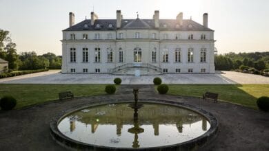 Un grand manoir, rappelant Châteaubriant, se distingue par son architecture symétrique et son jardin spacieux. Une fontaine ronde et réfléchissante orne le premier plan, entourée de pelouses et de bancs soigneusement entretenus. Le soleil se couche derrière la maison et projette une douce lueur sur cette scène pittoresque vue sur Actu44.fr.