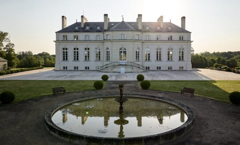 Un grand manoir, rappelant Châteaubriant, se distingue par son architecture symétrique et son jardin spacieux. Une fontaine ronde et réfléchissante orne le premier plan, entourée de pelouses et de bancs soigneusement entretenus. Le soleil se couche derrière la maison et projette une douce lueur sur cette scène pittoresque vue sur Actu44.fr.