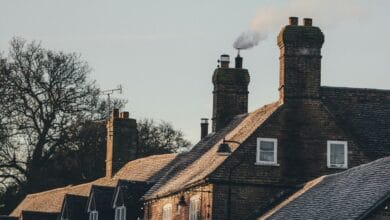 Une rangée de maisons en briques avec cheminées, dont l'une dégage de la fumée, évoque une matinée sereine à Châteaubriant. Les toits sont légèrement recouverts de givre tandis que les arbres nus se dressent sur un ciel clair de Loire-Atlantique, soulignant le charme tranquille de la région.