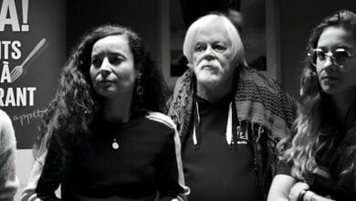 Une photo en noir et blanc montre trois personnes debout côte à côte. À gauche, une femme aux cheveux longs et bouclés, au centre, Paul Watson, un homme plus âgé aux cheveux blancs et à la barbe, et à droite, une femme portant des lunettes et aux cheveux longs. Une affiche de Sea Shepherd est partiellement visible à l'arrière-plan.