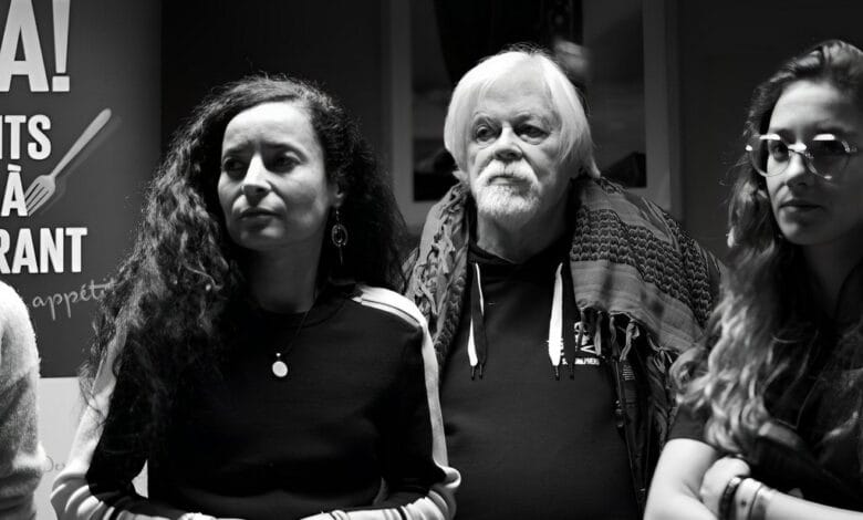 Une photo en noir et blanc montre trois personnes debout côte à côte. À gauche, une femme aux cheveux longs et bouclés, au centre, Paul Watson, un homme plus âgé aux cheveux blancs et à la barbe, et à droite, une femme portant des lunettes et aux cheveux longs. Une affiche de Sea Shepherd est partiellement visible à l'arrière-plan.