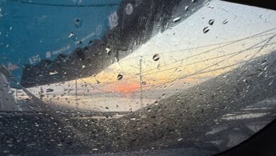 Des gouttes de pluie tombent sur un hublot d'un IMOCA avec vue sur un coucher de soleil envoûtant, rappelant la beauté sereine du Vendée Globe. Le ciel a des nuances d'orange et de bleu, visibles à travers la vitre mouillée, où les mâts d'un voilier et une partie d'une coque sont vaguement visibles au loin.