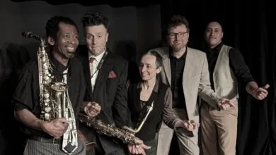 Un groupe de cinq musiciens nantais posent ensemble, tous souriants. Deux d'entre eux tiennent un saxophone, tandis que les autres se tiennent près les uns des autres, vêtus de tenues formelles ou semi-formelles. Le fond sombre évoque une scène ou un décor de spectacle en Loire-Atlantique.