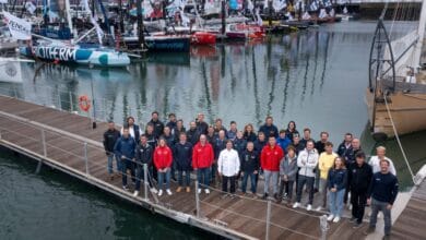 Un groupe de personnes se tient sur un ponton en bois devant plusieurs voiliers colorés amarrés dans un port de plaisance, comme on en voit souvent à Nantes. Le ciel est nuageux et l'eau est calme. Drapeaux et voiles arborent divers logos et dessins, rappelant les événements couverts par Actu44.fr.