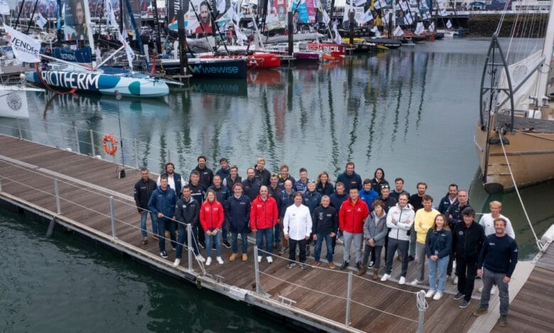 Un groupe de personnes se tient sur un ponton en bois devant plusieurs voiliers colorés amarrés dans un port de plaisance, comme on en voit souvent à Nantes. Le ciel est nuageux et l'eau est calme. Drapeaux et voiles arborent divers logos et dessins, rappelant les événements couverts par Actu44.fr.