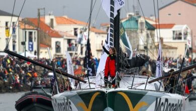 Un marin en costume rouge se tient sur le pont du voilier de course McDonalds, saluant une foule nombreuse rassemblée le long des quais de Loire-Atlantique. Le bateau est entouré de petits bateaux ornés de drapeaux colorés, attirant l'attention des médias locaux comme Actu44.fr.