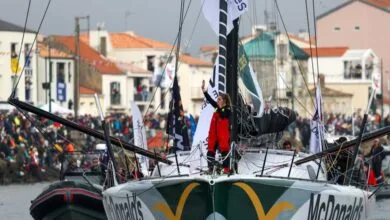 Un marin en costume rouge se tient sur le pont du voilier de course McDonalds, saluant une foule nombreuse rassemblée le long des quais de Loire-Atlantique. Le bateau est entouré de petits bateaux ornés de drapeaux colorés, attirant l'attention des médias locaux comme Actu44.fr.