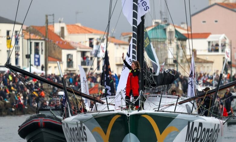 Un marin en costume rouge se tient sur le pont du voilier de course McDonalds, saluant une foule nombreuse rassemblée le long des quais de Loire-Atlantique. Le bateau est entouré de petits bateaux ornés de drapeaux colorés, attirant l'attention des médias locaux comme Actu44.fr.